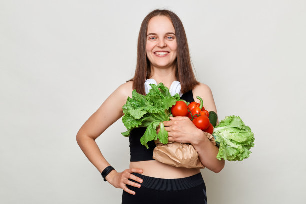自信的女性穿着运动服，拿着生菜、白菜、辣椒、牛油果，手放在臀部，看着镜头，宣传健康饮食。图片下载
