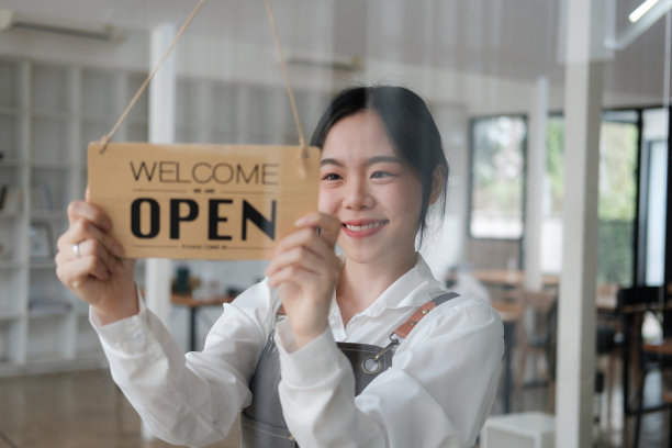 美丽而微笑的咖啡师，在她的咖啡馆前面有开放的标志，用美丽的微笑欢迎顾客来到她的店里。图片下载
