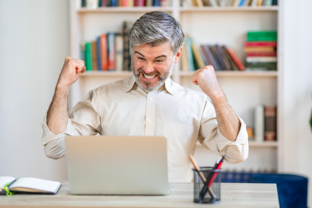 开朗的现代商人坐在办公空间的办公桌前，看着笔记本电脑屏幕高兴地尖叫，对交易的结果感到满意。高兴的人赢得了游戏，彩票。胜利的概念图片下载