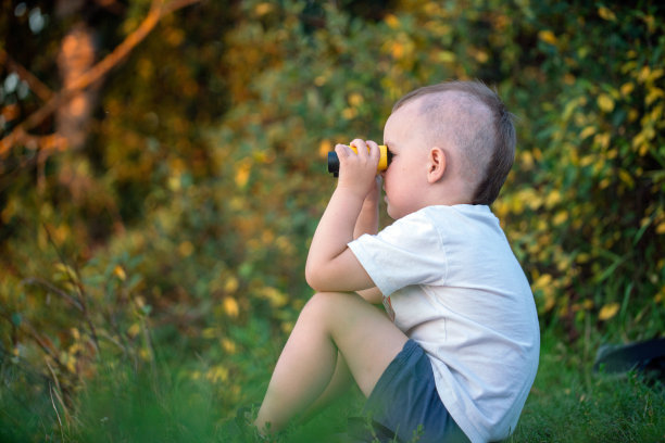 可爱的男孩坐在草地上用双筒望远镜看，侧视图图片下载