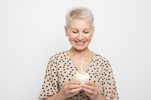 一位剪着短发的快乐老妇人手里拿着一个纸杯蛋糕，手里拿着蜡烛，一边庆祝生日一边笑着图片下载