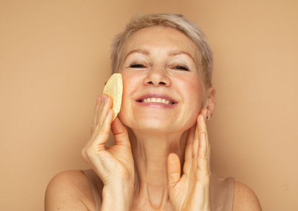 照片中善良的老妇人用海绵在米色背景上卸妆图片下载