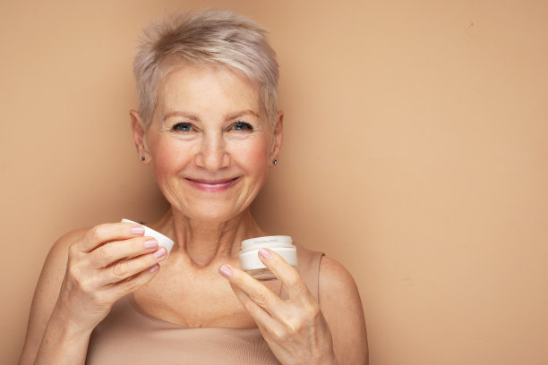 面带微笑的60多岁中年成熟女性看着镜头往脸上抹紧致面霜。图片下载