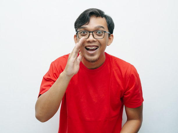 年轻的亚洲男子微笑着把双手放在嘴前低语的手势。红色t恤为8月17日印尼独立日概念图片下载