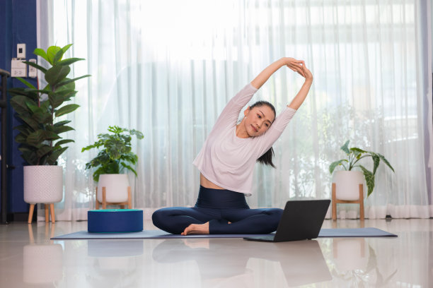 穿着白衬衫的亚洲女性在家里用笔记本电脑上练习瑜伽。图片下载