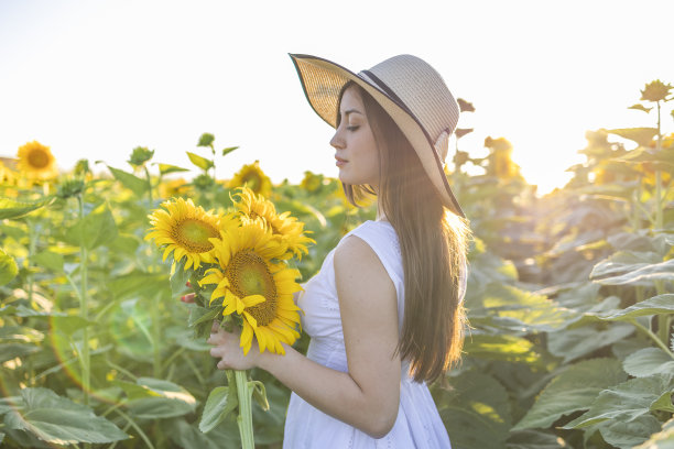 可爱的女孩拿着一束向日葵，在春天的向日葵地里摆姿势，穿着白色的裙子，头上戴着草帽。图片下载