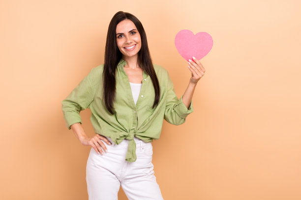 肖像的好心情少女的女人，长发型，穿绿色衬衫，手握心脏明信片孤立的米色背景图片下载
