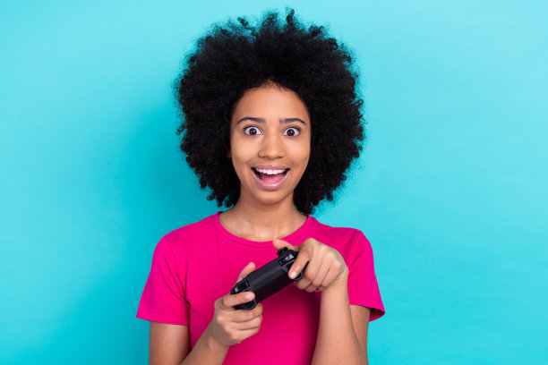 照片中，印象深刻的欣喜若狂的孩子穿着粉红色的t恤，手持操纵杆，在蓝色的背景上玩视频游戏图片下载