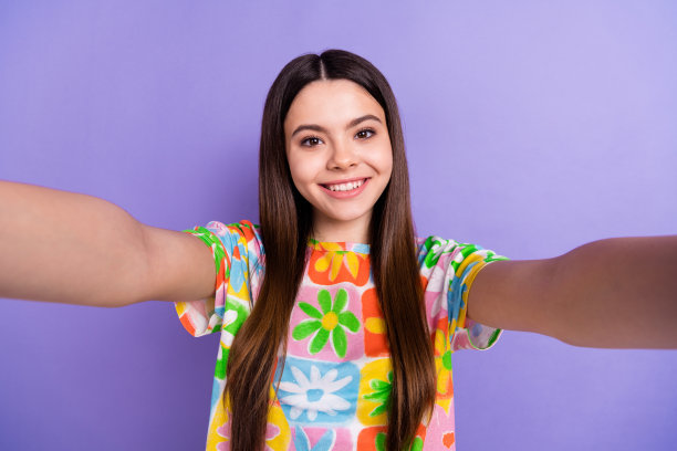 照片中，开朗满意的可爱女孩留着长发，穿着花t恤，在紫罗兰色的背景下自拍图片下载