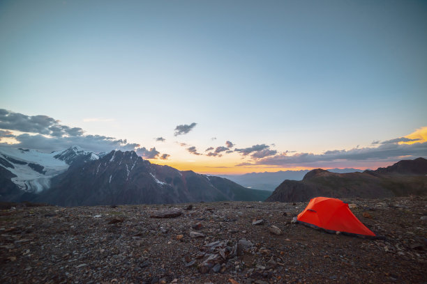 风景优美的高山景观，帐篷位于非常高的海拔，在橙色的黎明天空中可以看到大山。鲜艳的橙色帐篷，在多云的天空下，在日落的颜色下，可以看到令人敬畏的高山。图片下载