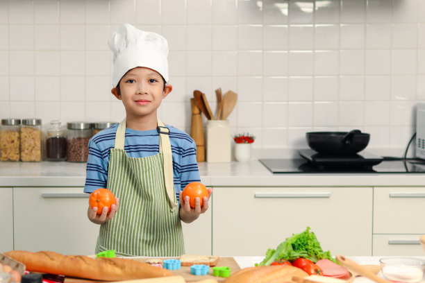开心微笑的亚洲男孩，系着围裙，戴着厨师帽，站在厨房里拿着橙色的水果，可爱的孩子厨师喜欢自己做饭，孩子喜欢吃水果。图片下载