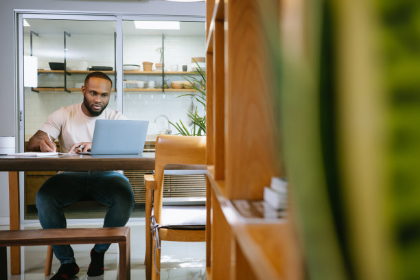非裔美国自由职业者在家用笔记本电脑工作，在厨房的桌子上处理文件。快乐的黑人男子在社交网络上交流，在线搜索信息。图片下载