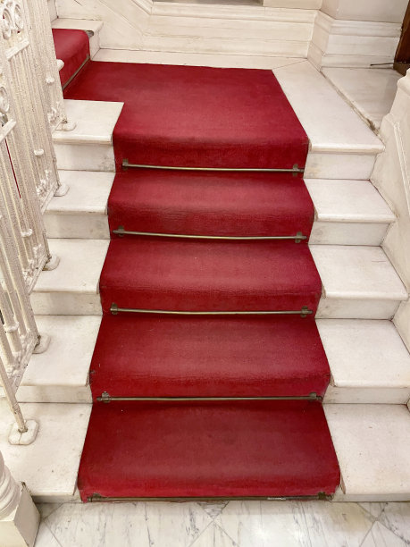 抛光白色的踏步和立管的特写图像，卷曲的石头楼梯，金属加工纺锤栏杆，红色天鹅绒地毯滑道与黄铜地毯杆连接，家居室内概念图片下载