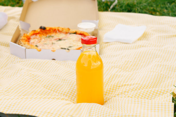 装在玻璃瓶里的橙汁模型，背景是黄色野餐垫上的披萨。天然多种维生素、桃子、果汁的生产概念及其用途。图片下载