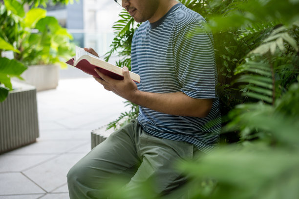 一个亚洲人坐在户外公园看书。年轻人拿着口袋书翻页。在大自然的背景下放松。图片下载