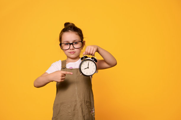可爱的小学生指着一个黑色的闹钟，显示上学时间，孤立的橙色背景图片下载