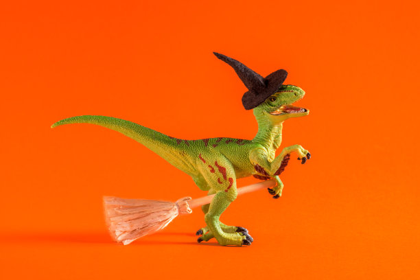 快乐的绿色玩具恐龙与女巫帽和扫帚在橙色背景。万圣节贺卡创意。图片下载