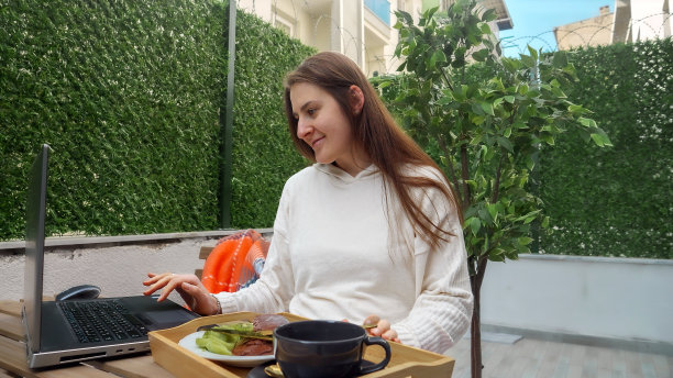 快乐微笑的女人端着托盘上的早餐走向露台，坐在桌子旁，用笔记本电脑工作。自由职业者的概念，在家工作，远程工作，在户外吃饭。图片下载