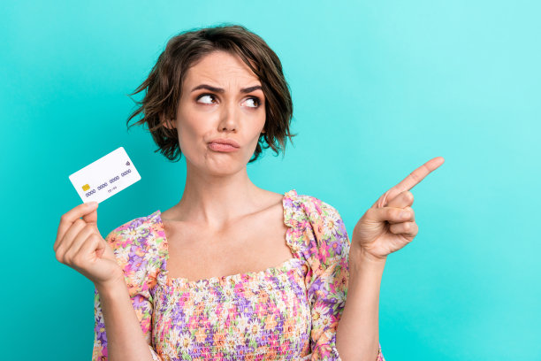 沉思沉思的女人穿着五颜六色的上衣，手持信用卡，在绿松石色的背景上看着空旷的空间图片下载