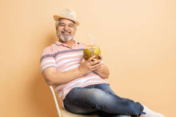 退休的印度高级男子穿着休闲的衣服和帽子，坐在椅子上喝着椰子水，在米色的工作室背景下放松。炎热的夏季，退休的生活，拷贝空间。图片下载