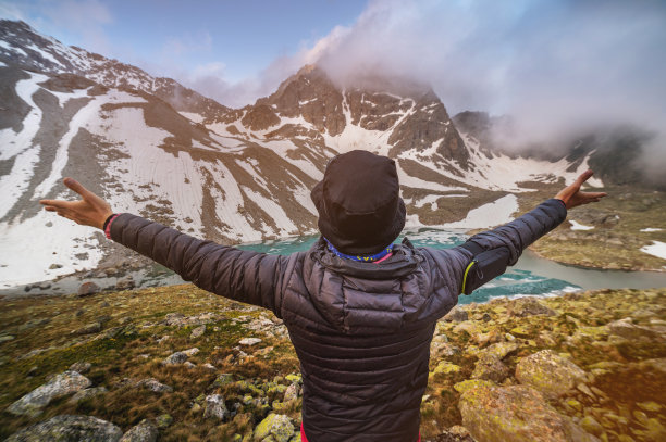 一个年轻人在山上的湖面上举起双手。不知名的游客站在岩石山顶，举起拳头，在白天多云的天空下欣赏山和湖的美景图片下载