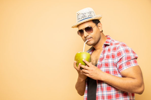 开朗的年轻印度男子戴着帽子和太阳镜，手里拿着椰子水，孤立在米色的工作室背景下。夏季饮品概念。图片下载