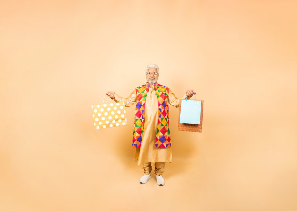 快乐的印度老人穿着传统的kurta服装，拿着购物袋，孤立地站在米色的工作室背景上。排灯节庆祝活动和节日销售概念。图片下载