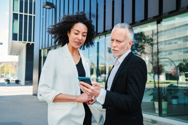 穿着西装的英俊商人站在公司附近的办公楼里，用智能手机向他的女同事展示信息。一对高管正在看手机图片下载