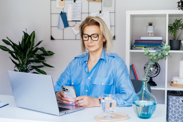 肖像50的自信成熟的女商人，中年经验丰富的高级女性专业人士使用电话，在舒适的办公室笔记本电脑上工作。女性CEO，创意企业家管理企业。图片下载