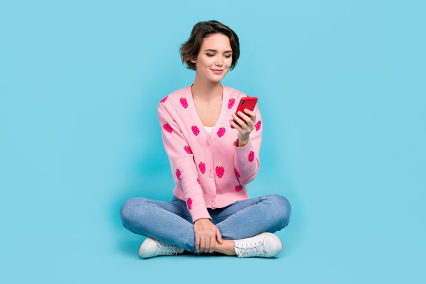 华丽的正面女士的全身照片坐在地板上交叉双腿使用智能手机聊天孤立的蓝色背景图片下载