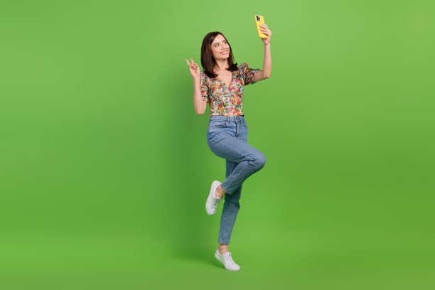 迷人友好的女士全身肖像拿着智能手机自拍，在绿色背景上显示v字图片下载