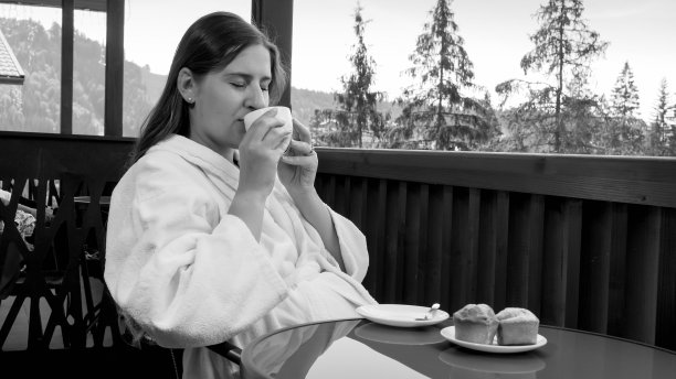 穿着浴袍的年轻女子坐在阳台上吃早餐的黑白肖像。人们在夏季到山区旅游度假的概念图片下载