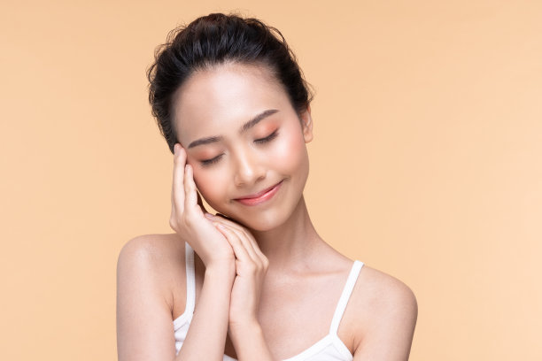 美丽的亚洲年轻女子抚摸柔软的脸颊，微笑，干净新鲜的皮肤，幸福和开朗，积极的情绪，隔离在米色背景，美容化妆品和面部护理概念图片下载