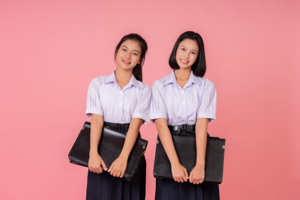 两个十几岁的亚洲女学生，短发和长发，笑容可爱，穿着校服。抱着两个黑色的包，身体，在粉红色的背景工作室拍摄。图片下载