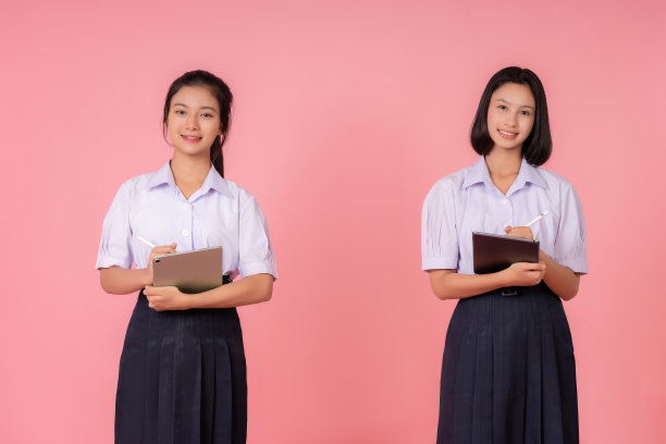 照片中的两个十几岁的亚洲女学生，短发和长发。穿着校服拿着笔记本和笔做作业，微笑的可爱女孩，在粉色背景的工作室里拍照图片下载
