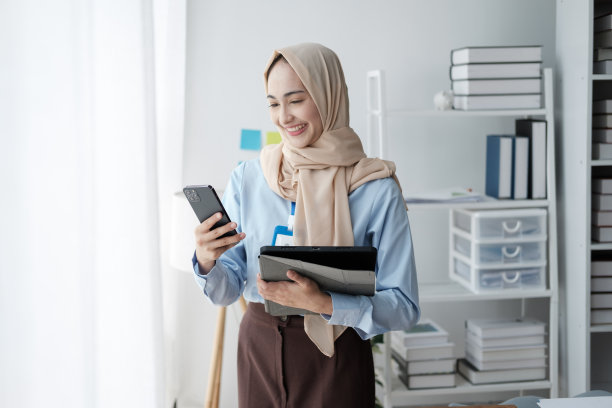 年轻漂亮的现代穆斯林妇女戴着头巾在办公室用笔记本电脑工作，在线教育，远程自由职业者，使用智能手机图片下载