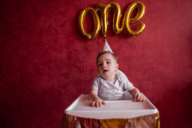 一岁大的小男孩戴着节日帽坐在儿童椅上，手里拿着布旗。首页图片下载