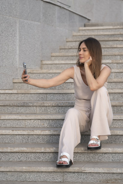 一名年轻女子坐在外面的楼梯台阶上，用她的智能手机自拍。图片下载