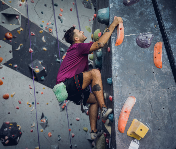 一位强壮的男性登山者用彩色的握把和绳索爬上一堵人造墙。图片下载