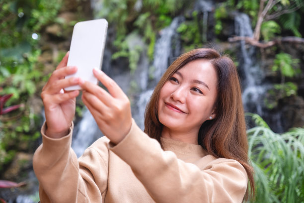 一位美丽的年轻亚洲女子在瀑布花园用手机自拍的肖像图片下载