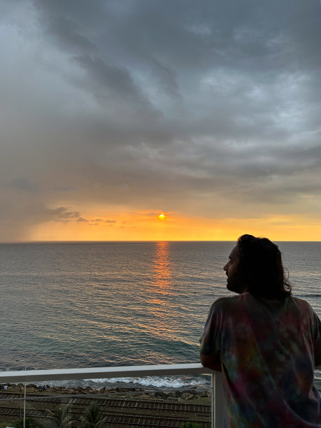 斯里兰卡，科伦坡，一名印度男子站在有玻璃安全屏障的酒店阳台上，倚在扶手上看橙色日落的天空和大海，忧郁的灰色云岸，水中反射的太阳图片下载