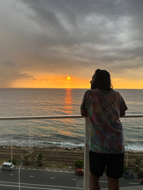 斯里兰卡，科伦坡，一名印度男子站在有玻璃安全屏障的酒店阳台上，靠在扶手上，看着橙色的夕阳天空和大海，忧郁的灰色云岸，水中反射的太阳图片下载