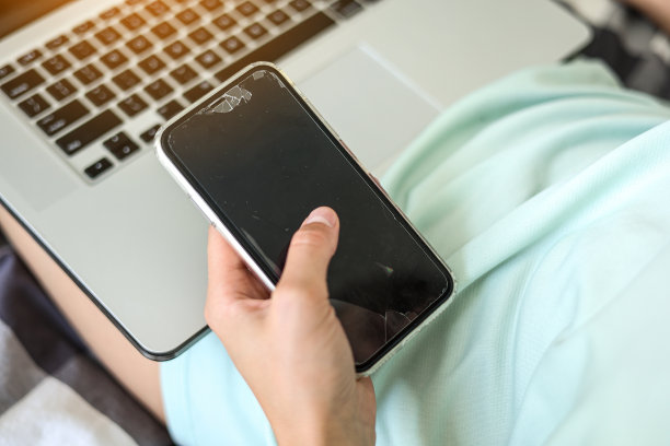 一个面目全非的女人在用手机发短信，手机的触摸屏坏了，玻璃也裂了。黑色的模型。笔记本电脑在后台。更换手机的碎玻璃。图片下载