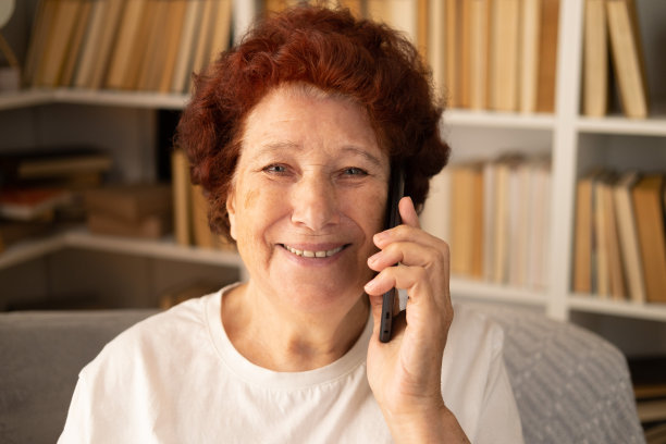 一个现代祖母的特写，她坐在书柜背景的扶手椅上，讲着电话，看着镜头，笑得很漂亮，牙齿洁白。图片下载