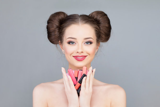 一个开朗的年轻成年女子，涂着亮粉色的唇膏，手里拿着彩色的口红，看着灰色背景上的相机图片下载