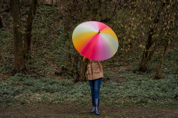 穿着米色外套和雨靴的年轻女子站在森林或公园里，手持色彩鲜艳的彩虹伞。女孩旋转雨伞以获得模糊效果。慢速快门。城市的秋天天气图片下载
