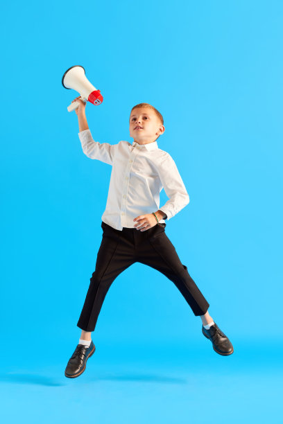 小男孩，穿着白衬衫和黑裤子的孩子，扩音器在蓝色的工作室背景下跳跃图片下载
