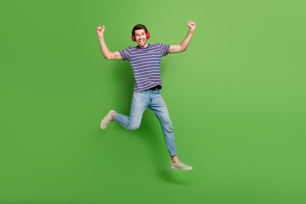 全身照片正面幸运的家伙穿条纹t恤跳高听音乐上升的拳头孤立的绿色背景图片下载