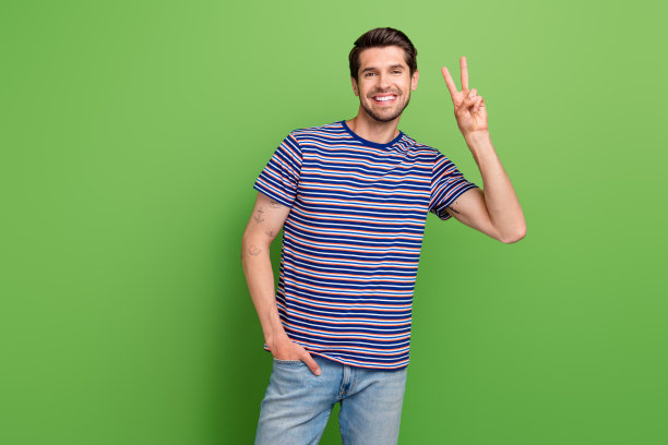 心情好的男人穿着条纹时尚的衣服，手臂放在口袋里，在绿色的背景上显示v字手势打招呼图片下载