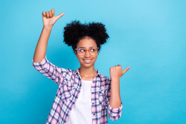 照片中兴奋积极的女孩穿着格子衬衫，戴着眼镜指着两个拇指空白的空间孤立的蓝色背景图片下载
